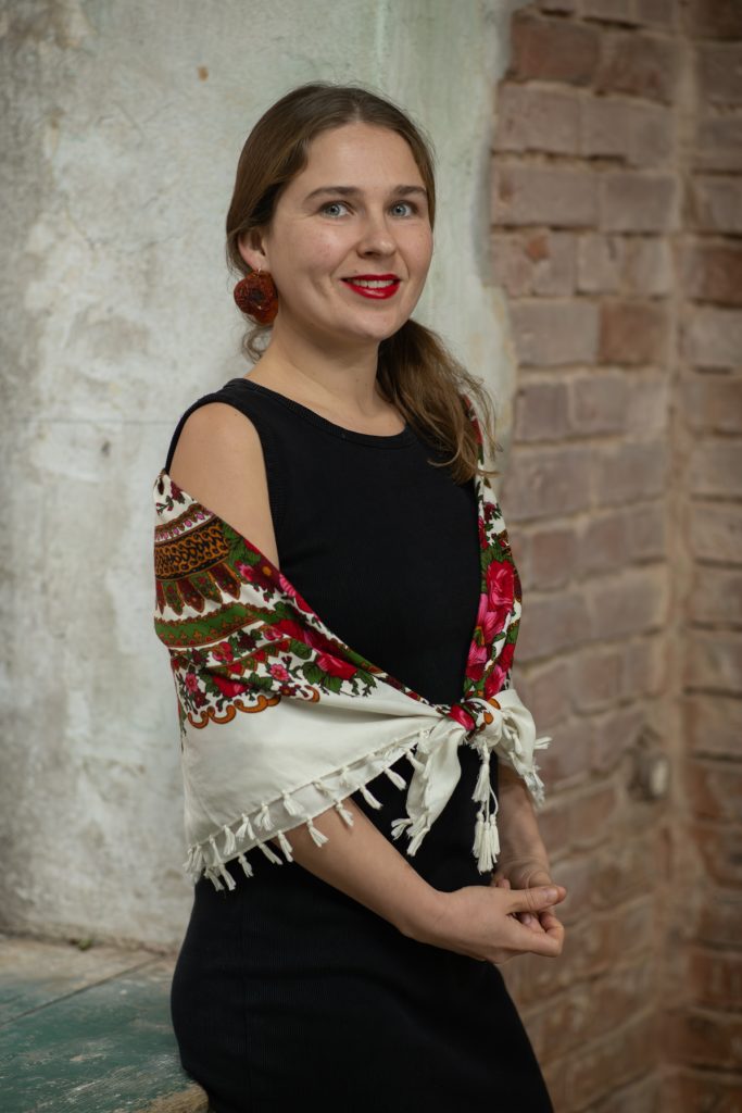 Portret Anastazji z Mariupola w półprofilu z uśmiechem, w kwiecistej chuście i czerwonej szmince