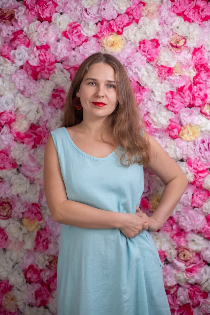 Anastazja z Mariupola w błękitnej sukni na tle ściany pokrytej różowymi i białymi kwiatami